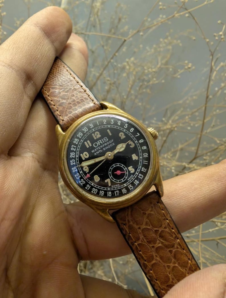 Puede incluir: Reloj Oris vintage dorado con esfera negra, n&uacute;meros y agujas blancos. El reloj tiene una correa de cuero marr&oacute;n. La esfera del reloj incluye el texto "ORIS WATERPROOF ANTI-SHOCK".
