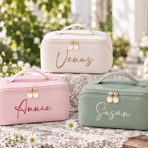 May include: Three rectangular cosmetic bags with top handles and gold-toned double zipper pulls. The white bag features the name Venus, the pink bag features the name Annie, and the sage green bag features the name Susan in cursive script.