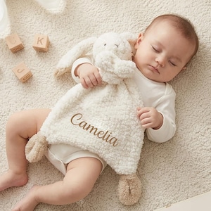 May include: A cream-colored plush security blanket featuring a stuffed rabbit head with long ears and tan accents on the paws and ears. The soft, textured fabric is embroidered with the name Camelia in brown script lettering on the front.