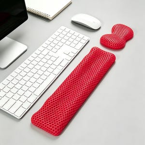 May include: A computer setup featuring a white keyboard, mouse, and a red honeycomb-patterned wrist rest and mouse pad. The wrist rest is long and rectangular, while the mouse pad is shaped to fit the wrist. The items are on a gray surface.