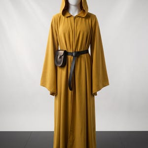 Pode incluir: Uma túnica longa até ao chão em amarelo mostarda com capuz, mangas compridas e largas, e fecho de correr frontal. Um cinto fino de couro preto está ajustado à cintura, com uma pequena bolsa de couro preta estruturada presa ao lado.