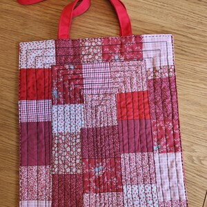 Può includere: Borsa tote fatta a mano con un design patchwork nei toni del rosso e del rosa. La borsa presenta una varietà di tessuti floreali e fantasia, con un manico rosso pieno. La borsa è trapuntata con filo nero.