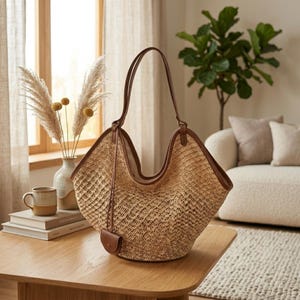 May include: A woven straw tote bag with brown leather trim and handles. The bag has a distinctive shape and a small matching pouch. The bag is on a wooden table, with a neutral background.