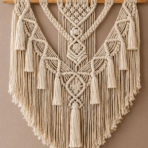 Op de afbeelding: Een crèmekleurige macramé wandhanger opgehangen aan een horizontale houten stok. Het stuk bevat ingewikkelde knooppatronen, waaronder ruitvormen en verticale franjes, afgewerkt met meerdere decoratieve kwastjes langs de randen.