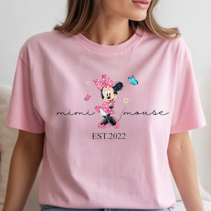 May include: A pale pink t-shirt with a Minnie Mouse design, including a pink bow and a blue butterfly. The words "mimi mouse" are in cursive, with "EST.2022" underneath. The shirt is worn with blue jeans.