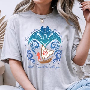 Op de afbeelding: Lichtgrijs t-shirt met een kleurrijke afbeelding van een boot die op blauwe golven vaart, met een silhouet van een persoon. De tekst luidt: "Er is geen plek waar je naartoe kunt gaan waar ik niet bij je zal zijn."