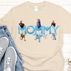 May include: Beige t-shirt with the word "MOMMY" in blue, featuring characters from the movie Frozen. The shirt includes images of Anna, Elsa, Kristoff, Sven, Olaf, and Marshmallow. The shirt is paired with blue jeans and gray sneakers.