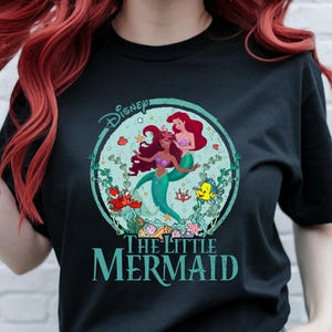 K&ouml;nnte beinhalten: Schwarzes T-Shirt mit einem farbenfrohen Grafikdesign von Arielle und einer weiteren Meerjungfrau aus Disneys Arielle, die Meerjungfrau. Das Design befindet sich in einem t&uuml;rkisfarbenen Kreis mit Wasserpflanzen und Meeresbewohnern. Das Disney-Logo und der Filmtitel sind ebenfalls zu sehen.