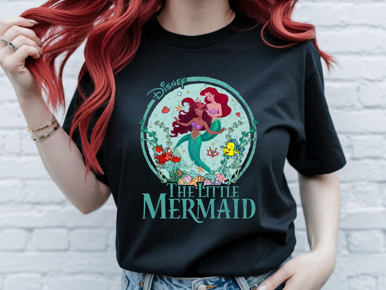 K&ouml;nnte beinhalten: Schwarzes T-Shirt mit einem farbenfrohen Grafikdesign von Arielle und einer weiteren Meerjungfrau aus Disneys Arielle, die Meerjungfrau. Das Design befindet sich in einem t&uuml;rkisfarbenen Kreis mit Wasserpflanzen und Meeresbewohnern. Das Disney-Logo und der Filmtitel sind ebenfalls zu sehen.