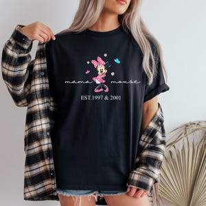 May include: Black t-shirt with a Minnie Mouse design in pink and white, accompanied by the words "mama mouse" and the years "EST.1997 & 2001". The design includes butterflies. The shirt is worn with a plaid flannel.
