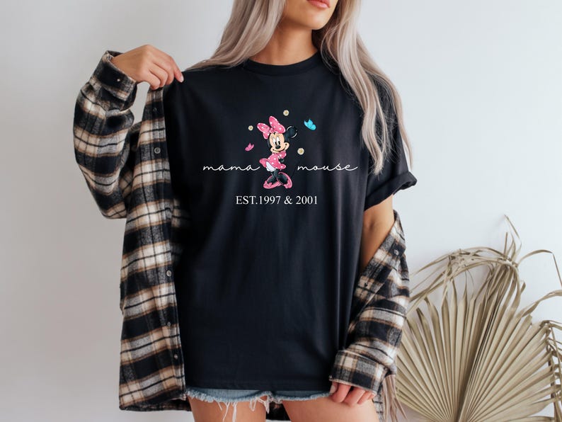 May include: Black t-shirt with a Minnie Mouse design in pink and white, accompanied by the words "mama mouse" and the years "EST.1997 & 2001". The design includes butterflies. The shirt is worn with a plaid flannel.