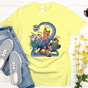 Puede incluir: Una camiseta amarilla clara con una ilustraci&oacute;n de dibujos animados de personajes de animales, incluyendo un zorro, un conejo y un castor, envueltos alrededor de una serpiente azul. Los nombres de los personajes est&aacute;n escritos en la camiseta. La camiseta se combina con vaqueros azules y zapatillas grises.