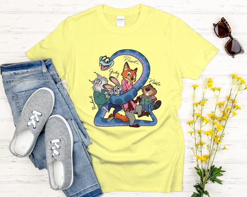 Puede incluir: Una camiseta amarilla clara con una ilustraci&oacute;n de dibujos animados de personajes de animales, incluyendo un zorro, un conejo y un castor, envueltos alrededor de una serpiente azul. Los nombres de los personajes est&aacute;n escritos en la camiseta. La camiseta se combina con vaqueros azules y zapatillas grises.
