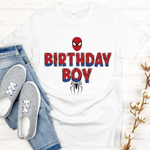 Może przedstawiać: Biała koszulka z napisem "BIRTHDAY BOY" w kolorze czerwono-niebieskim, z grafiką Spider-Mana. Koszulka jest połączona z niebieskimi dżinsami i szarymi trampkami.