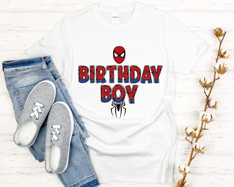 Może przedstawiać: Biała koszulka z napisem "BIRTHDAY BOY" w kolorze czerwono-niebieskim, z grafiką Spider-Mana. Koszulka jest połączona z niebieskimi dżinsami i szarymi trampkami.