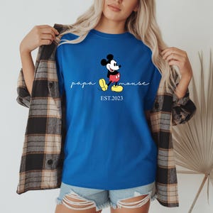 May include: Royal blue t-shirt with a Mickey Mouse cartoon design. The shirt features the words "papa mouse" in cursive and "EST.2023" below the character. Worn with denim shorts and a plaid shirt.