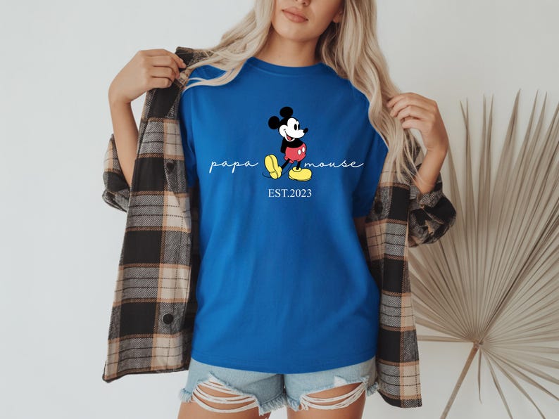 May include: Royal blue t-shirt with a Mickey Mouse cartoon design. The shirt features the words "papa mouse" in cursive and "EST.2023" below the character. Worn with denim shorts and a plaid shirt.
