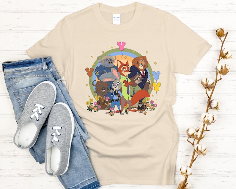 Peut inclure: T-shirt beige avec un motif de dessin anim&eacute; color&eacute; de personnages d'animaux, un arc-en-ciel, des fleurs et des ballons en forme de Mickey Mouse. Le t-shirt est associ&eacute; &agrave; un jean bleu et des baskets grises.