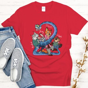 Puede incluir: Camiseta roja con una ilustraci&oacute;n de dibujos animados de personajes animales, incluyendo un zorro, un conejo y un castor, con el texto "2" y nombres de personajes. La camiseta se combina con vaqueros azules y zapatillas grises.