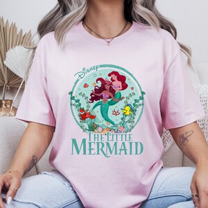 K&ouml;nnte beinhalten: Hellrosa T-Shirt mit einem kreisf&ouml;rmigen Aufdruck von Arielle und anderen Figuren aus Disneys The Little Mermaid. Der Aufdruck enth&auml;lt das Disney-Logo und die Worte "The Little Mermaid" in Gr&uuml;n. Das T-Shirt hat einen Rundhalsausschnitt.