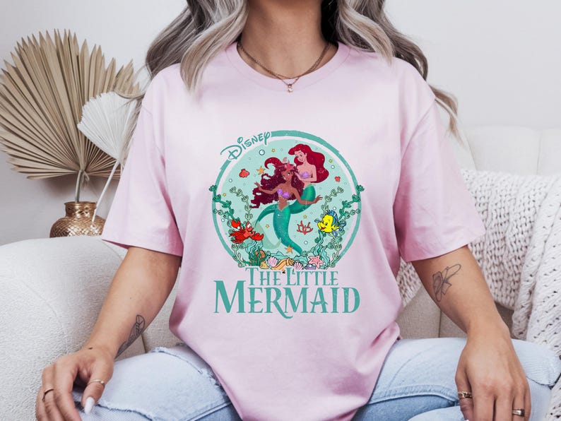 K&ouml;nnte beinhalten: Hellrosa T-Shirt mit einem kreisf&ouml;rmigen Aufdruck von Arielle und anderen Figuren aus Disneys The Little Mermaid. Der Aufdruck enth&auml;lt das Disney-Logo und die Worte "The Little Mermaid" in Gr&uuml;n. Das T-Shirt hat einen Rundhalsausschnitt.