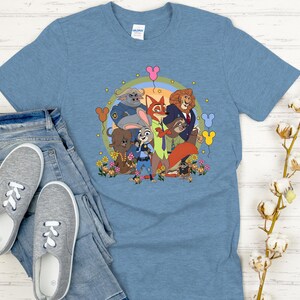 Peut inclure: T-shirt bleu clair avec un motif color&eacute; des personnages du film d'animation Zootopie. Le design comprend Judy Hopps, Nick Wilde et d'autres personnages, entour&eacute;s de fleurs et de ballons. Le t-shirt est associ&eacute; &agrave; un jean bleu et des baskets grises.