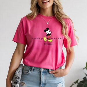 May include: A vibrant pink t-shirt with a Mickey Mouse graphic. The design includes the words "mickey mouse" in a script font and "EST.2024" below the character. The shirt is a short-sleeved crew neck.