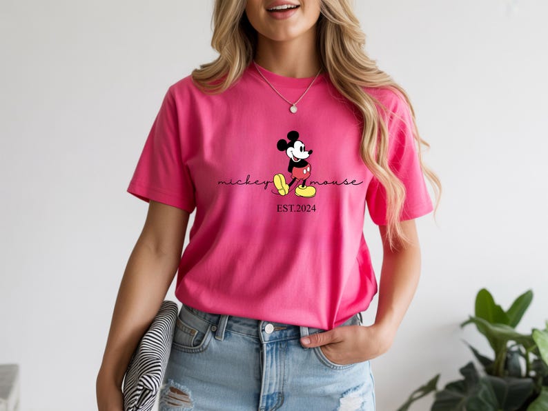 May include: A vibrant pink t-shirt with a Mickey Mouse graphic. The design includes the words "mickey mouse" in a script font and "EST.2024" below the character. The shirt is a short-sleeved crew neck.