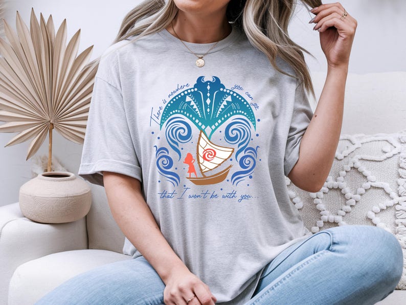 Op de afbeelding: Lichtgrijs t-shirt met een kleurrijke afbeelding van een boot die op blauwe golven vaart, met een silhouet van een persoon. De tekst luidt: "Er is geen plek waar je naartoe kunt gaan waar ik niet bij je zal zijn."