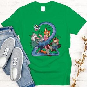 Puede incluir: Camiseta verde con personajes de dibujos animados de Zootopia. El dise&ntilde;o incluye a Judy, Nick y otros personajes, con el n&uacute;mero "2" y los nombres de los personajes. La camiseta se combina con vaqueros azules y zapatillas grises.