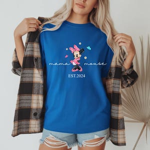May include: A royal blue t-shirt with a Minnie Mouse graphic, the text "mama mouse" and "EST.2024". The graphic shows Minnie Mouse in a pink dress and bow, with blue butterflies. The shirt is worn with denim shorts and a plaid shirt.