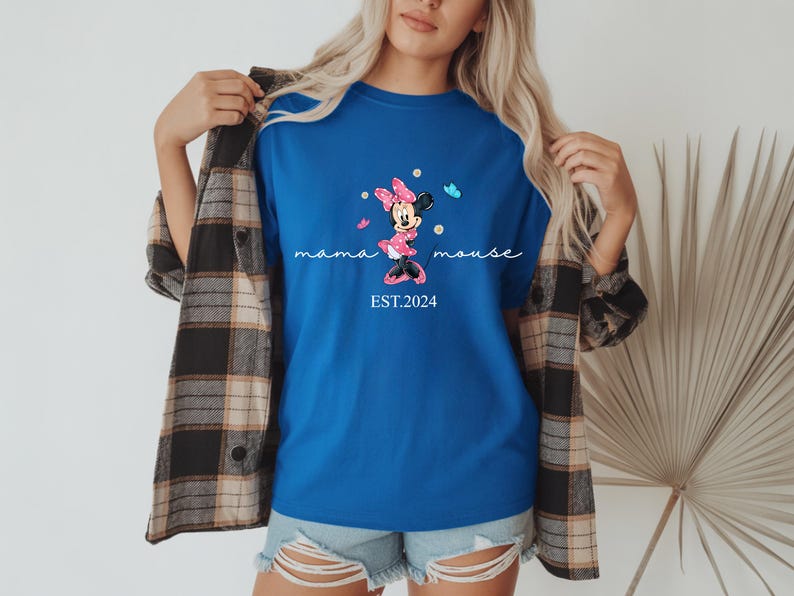 May include: A royal blue t-shirt with a Minnie Mouse graphic, the text "mama mouse" and "EST.2024". The graphic shows Minnie Mouse in a pink dress and bow, with blue butterflies. The shirt is worn with denim shorts and a plaid shirt.