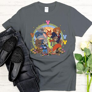 Peut inclure: Un t-shirt gris fonc&eacute; avec une illustration de dessin anim&eacute; color&eacute;e des personnages de Zootopie. Le design comprend Judy Hopps, Nick Wilde et d'autres personnages, entour&eacute;s de fleurs et de ballons. Le t-shirt est associ&eacute; &agrave; un jean et des chaussures noirs.