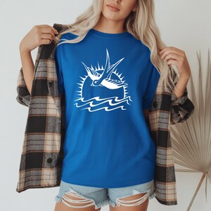 K&ouml;nnte beinhalten: Ein k&ouml;nigsblaues T-Shirt mit einer wei&szlig;en Grafik einer Schwalbe, die &uuml;ber Wellen und einer Sonne fliegt. Das Shirt wird mit Jeansshorts und einem karierten Flanellhemd getragen.