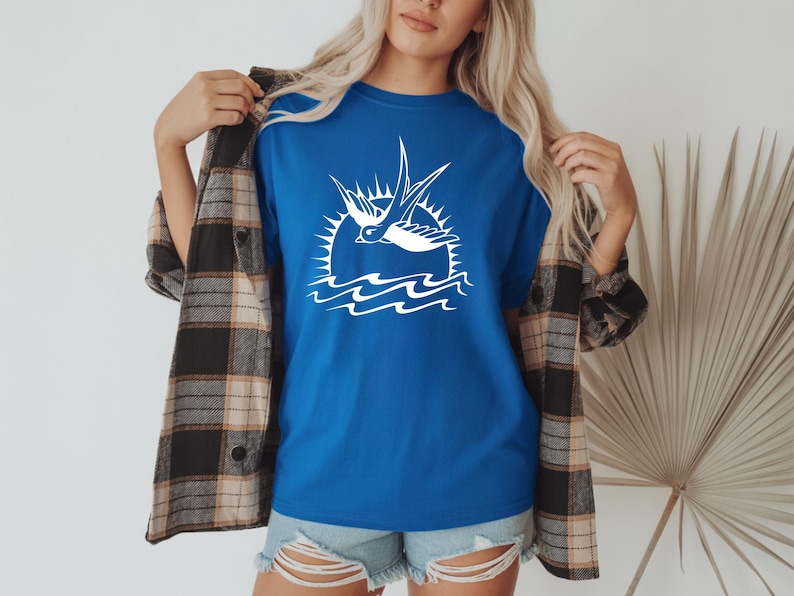 K&ouml;nnte beinhalten: Ein k&ouml;nigsblaues T-Shirt mit einer wei&szlig;en Grafik einer Schwalbe, die &uuml;ber Wellen und einer Sonne fliegt. Das Shirt wird mit Jeansshorts und einem karierten Flanellhemd getragen.