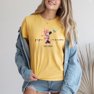 May include: Mustard yellow t-shirt with a Minnie Mouse design and the text "gigi mouse EST.2020". The shirt is paired with a denim jacket and shorts. The Minnie Mouse graphic includes a pink bow and a blue butterfly.