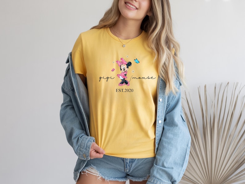 May include: Mustard yellow t-shirt with a Minnie Mouse design and the text "gigi mouse EST.2020". The shirt is paired with a denim jacket and shorts. The Minnie Mouse graphic includes a pink bow and a blue butterfly.