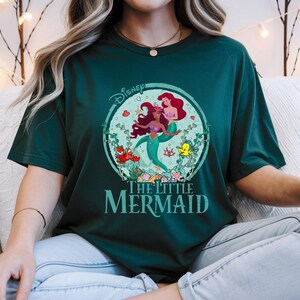 K&ouml;nnte beinhalten: Dunkelgr&uuml;nes T-Shirt mit einem kreisf&ouml;rmigen Aufdruck von Arielle und anderen Figuren aus Disneys Arielle, die Meerjungfrau. Die Grafik enth&auml;lt das Disney-Logo und die Worte "The Little Mermaid" in T&uuml;rkis. Das Shirt hat eine lockere Passform.