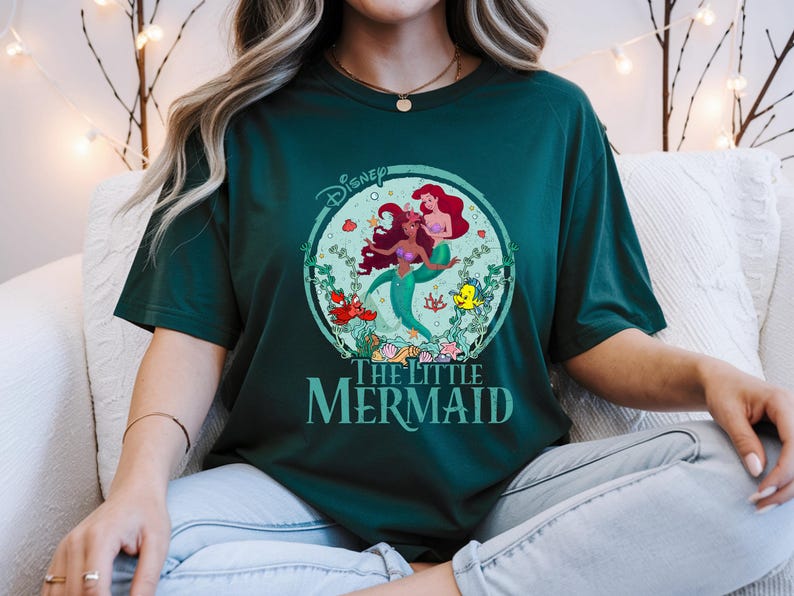 K&ouml;nnte beinhalten: Dunkelgr&uuml;nes T-Shirt mit einem kreisf&ouml;rmigen Aufdruck von Arielle und anderen Figuren aus Disneys Arielle, die Meerjungfrau. Die Grafik enth&auml;lt das Disney-Logo und die Worte "The Little Mermaid" in T&uuml;rkis. Das Shirt hat eine lockere Passform.
