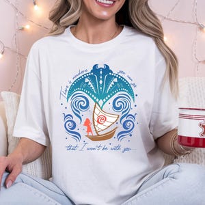 Op de afbeelding: Wit T-shirt met een kleurrijke afbeelding van een boot, golven en de tekst "There is nowhere you can go that I won't be with you...". Een rood-witte mok met een peperkoekmannetje wordt in de hand gehouden.