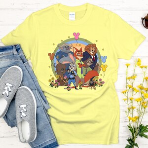 Peut inclure: Un t-shirt jaune clair avec une illustration de personnages de Zootopie. Le design comprend Judy Hopps, Nick Wilde et d'autres personnages, entour&eacute;s de fleurs et de ballons. Le t-shirt est associ&eacute; &agrave; un jean bleu et des baskets grises.