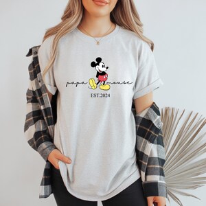 May include: Light grey t-shirt with a Mickey Mouse graphic, the words "papa mouse" and "EST.2024". The shirt has rolled sleeves and is worn with a black and white checked shirt.