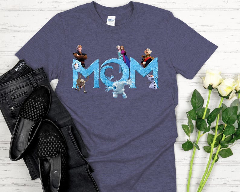 May include: A blue t-shirt featuring the word "MOM" in a stylized, icy blue font. The letters are decorated with characters from the movie Frozen. The shirt is paired with black jeans and shoes.