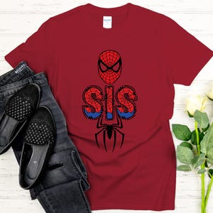 Może przedstawiać: Bordowa koszulka z grafiką Spider-Mana. Projekt zawiera maskę Spider-Mana, słowo "SIS" w czcionce przypominającej pajęczynę i sylwetkę pająka. Koszulka jest zestawiona z ciemnymi dżinsami i czarnymi balerinami.