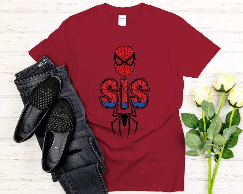 Może przedstawiać: Bordowa koszulka z grafiką Spider-Mana. Projekt zawiera maskę Spider-Mana, słowo "SIS" w czcionce przypominającej pajęczynę i sylwetkę pająka. Koszulka jest zestawiona z ciemnymi dżinsami i czarnymi balerinami.