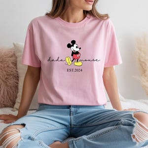 May include: A pale pink t-shirt with a Mickey Mouse graphic. The design includes the cursive text "dada mouse", with Mickey Mouse standing beside it. Below the text is "EST.2024". The shirt is made of a soft material.