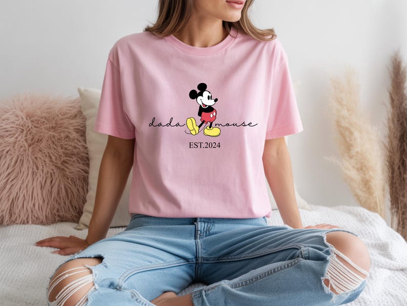 May include: A pale pink t-shirt with a Mickey Mouse graphic. The design includes the cursive text "dada mouse", with Mickey Mouse standing beside it. Below the text is "EST.2024". The shirt is made of a soft material.