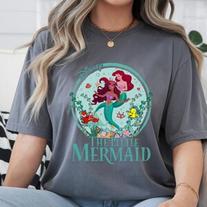 K&ouml;nnte beinhalten: Graues T-Shirt mit einem kreisf&ouml;rmigen Grafikdesign von Disneys "Arielle, die Meerjungfrau". Die Grafik zeigt Arielle, Fabius und Sebastian, umgeben von Wasserpflanzen. Der Text "Disney" und "The Little Mermaid" sind ebenfalls sichtbar.