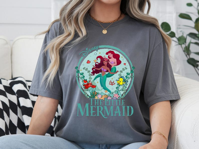 K&ouml;nnte beinhalten: Graues T-Shirt mit einem kreisf&ouml;rmigen Grafikdesign von Disneys "Arielle, die Meerjungfrau". Die Grafik zeigt Arielle, Fabius und Sebastian, umgeben von Wasserpflanzen. Der Text "Disney" und "The Little Mermaid" sind ebenfalls sichtbar.