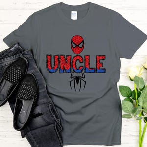 Może przedstawiać: Szara koszulka z motywem Spider-Mana. Słowo "UNCLE" jest wydrukowane w kolorze czerwonym i niebieskim, z maską Spider-Mana powyżej i grafiką pająka poniżej. Koszulka jest połączona z czarnymi dżinsami i butami.